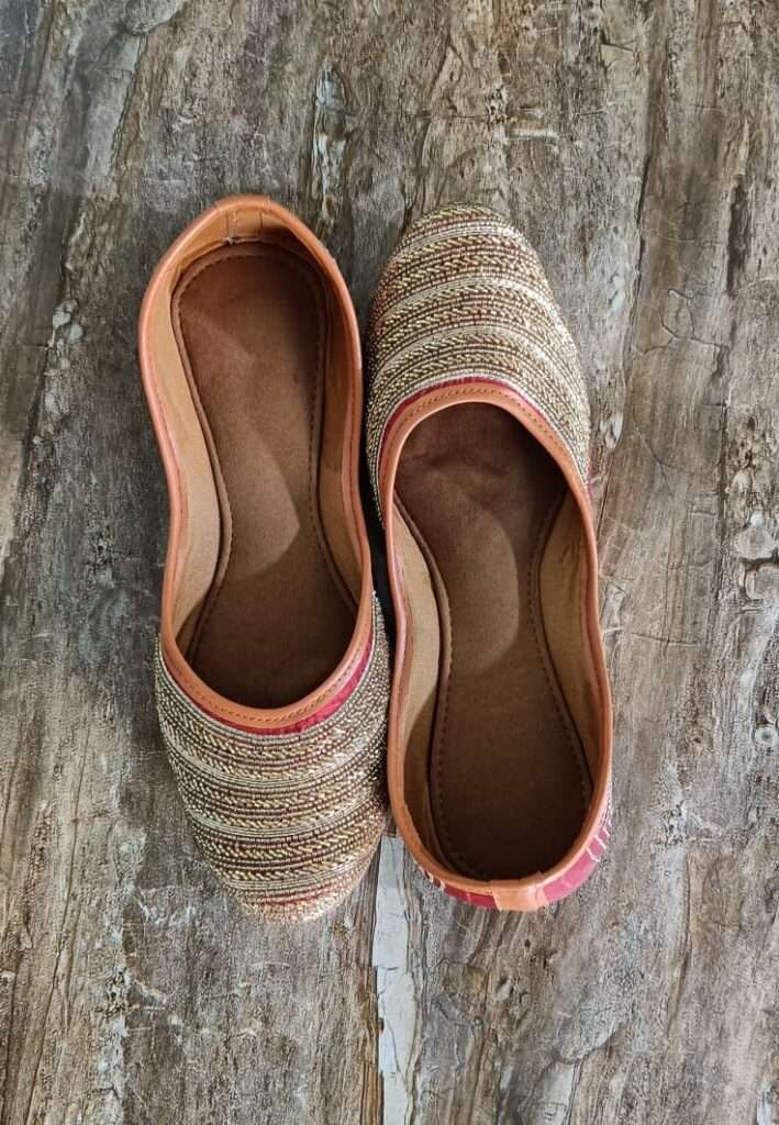 An image showcases exquisite ladies' Indian ethnic juttis. Maroon base, intricate gold zari embroidery in floral patterns. Pointed toe, shimmering gold border. A fusion of heritage and elegance.